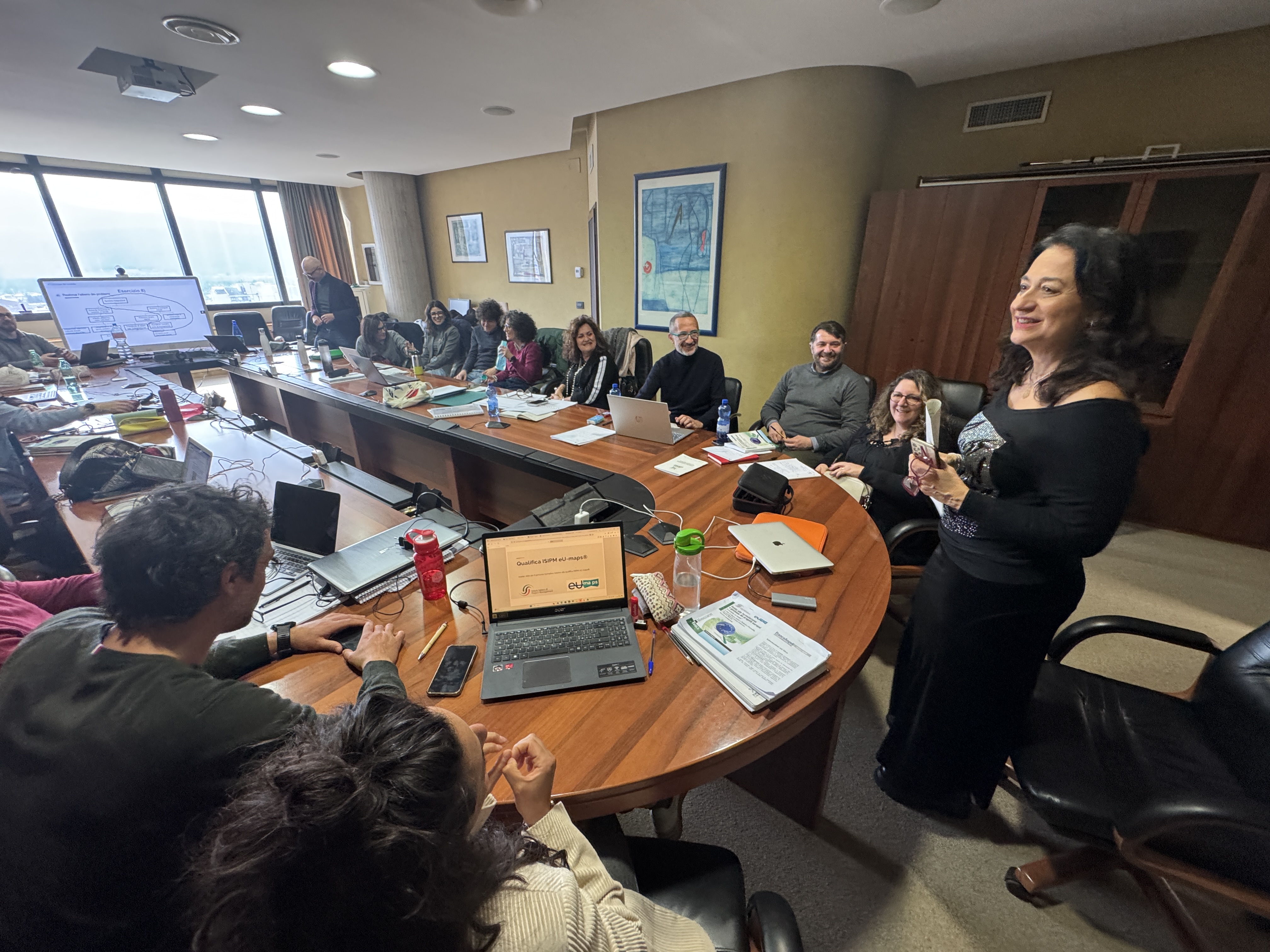 Donna in piedi presenta a un gruppo seduto attorno a un tavolo da riunione con laptop e documenti, in una sala luminosa.