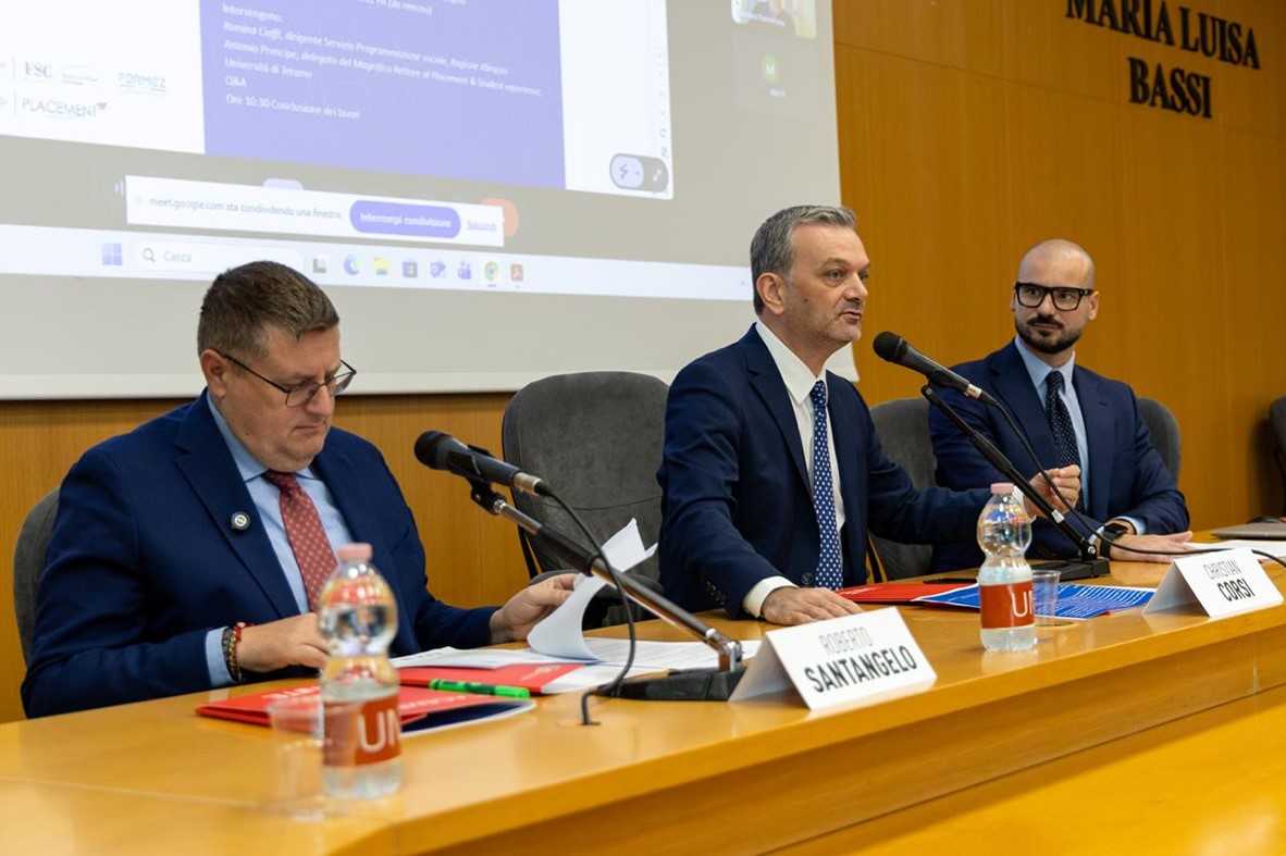 Foto dalla presentazione del progetto. Tre persone sedute ad una scrivania parlano al microfono, con dietro immagini proiettate
